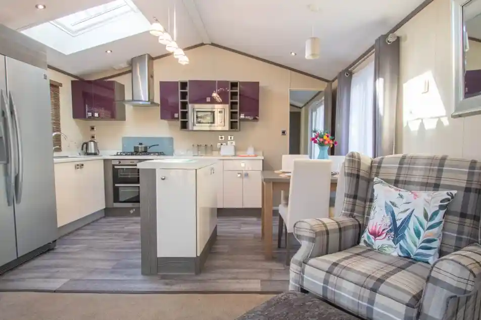 An open plan kitchen, dining, living room with Velux roof window and laminate wood flooring to the kitchen area. A mixture of white gloss and purple gloss cabinets, a kitchen island and a dining table with chairs separates the kitchen area from the lounge.