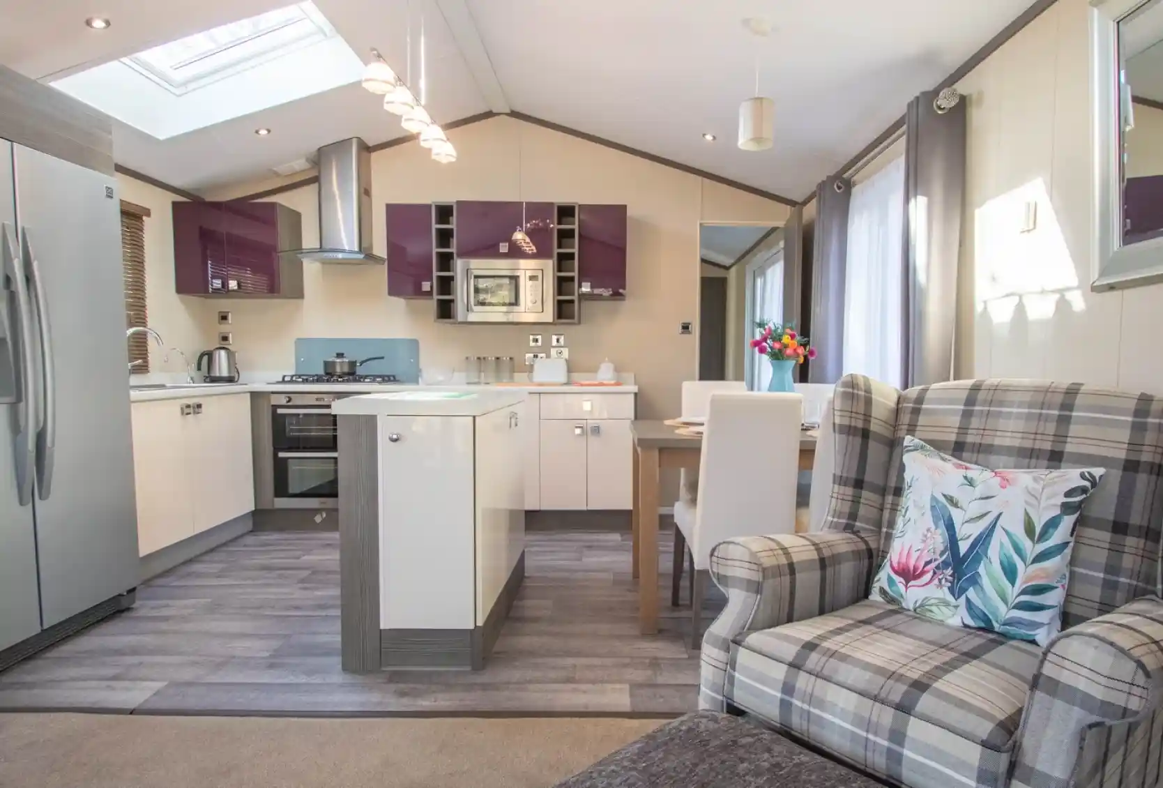 An open plan kitchen, dining, living room with Velux roof window and laminate wood flooring to the kitchen area. A mixture of white gloss and purple gloss cabinets, a kitchen island and a dining table with chairs separates the kitchen area from the lounge.