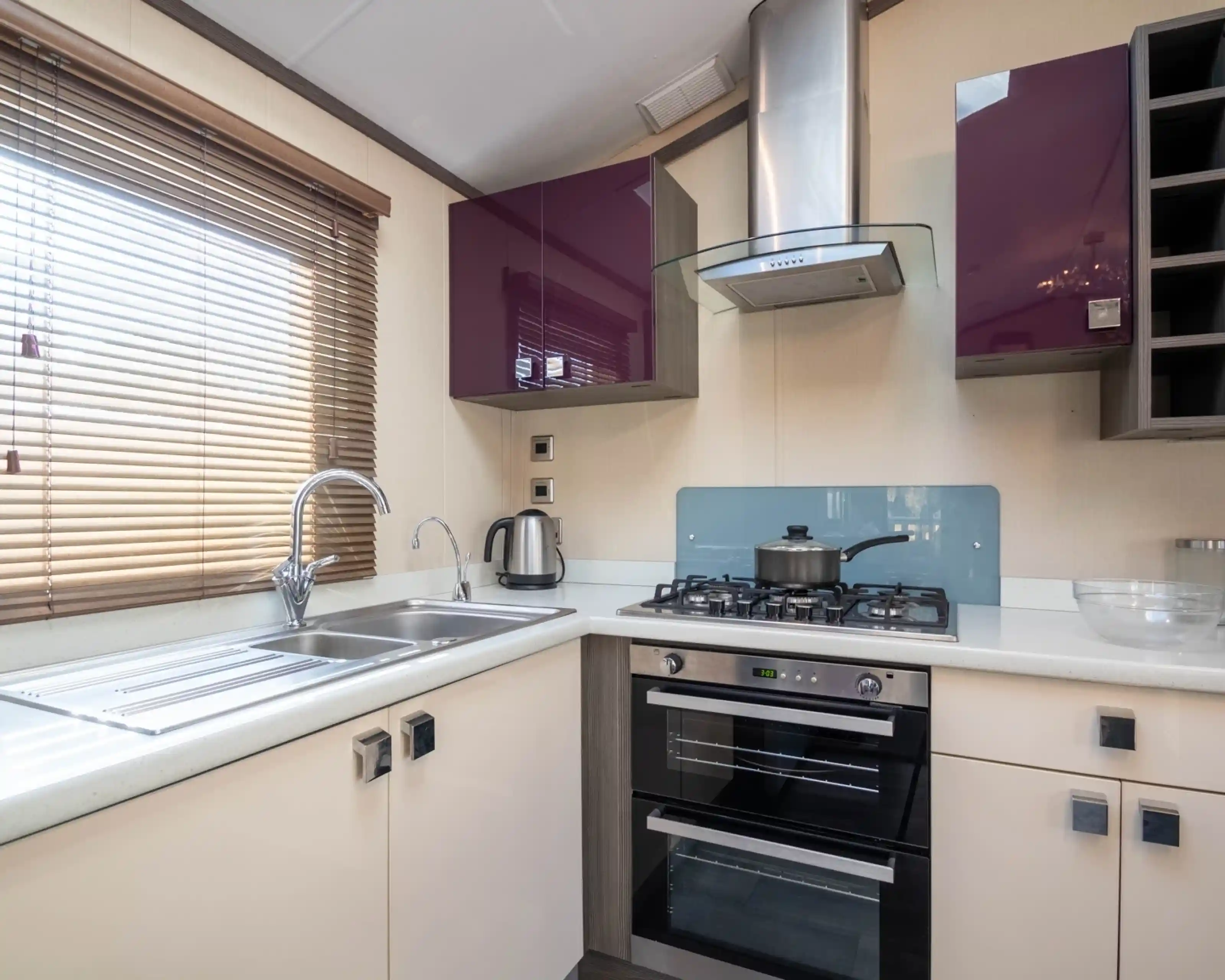 A bright small kitchen with purple and cream gloss cabinets, including an oven with gas hob, extractor hood and stainless steel one-and-a-half bowl sink unit, together with a window with blinds.