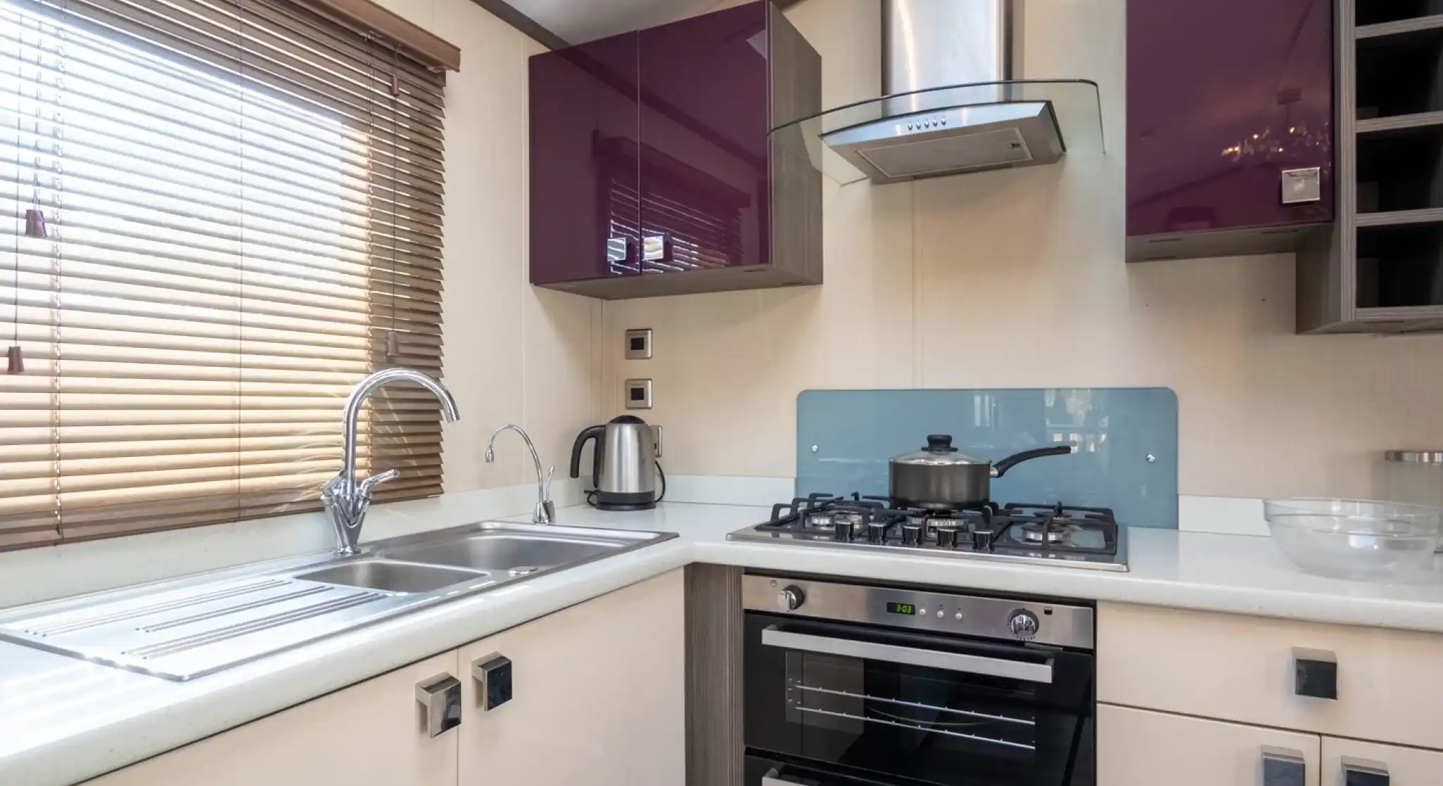A bright small kitchen with purple and cream gloss cabinets, including an oven with gas hob, extractor hood and stainless steel one-and-a-half bowl sink unit, together with a window with blinds.