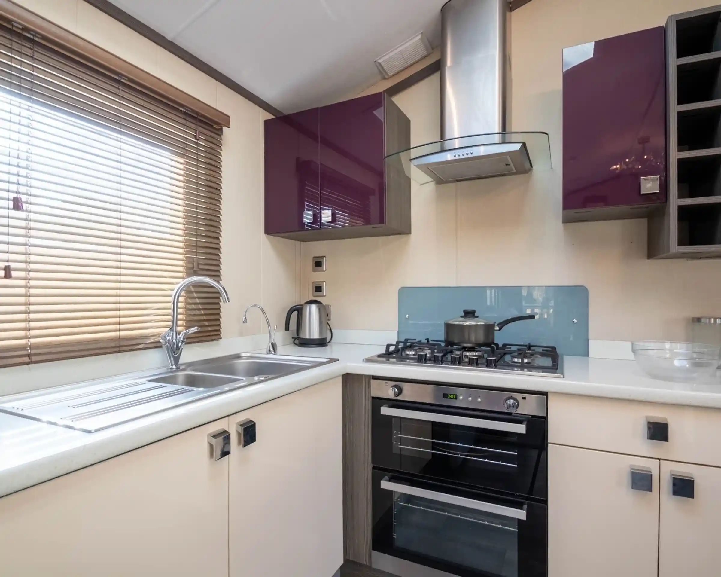 A bright small kitchen with purple and cream gloss cabinets, including an oven with gas hob, extractor hood and stainless steel one-and-a-half bowl sink unit, together with a window with blinds.