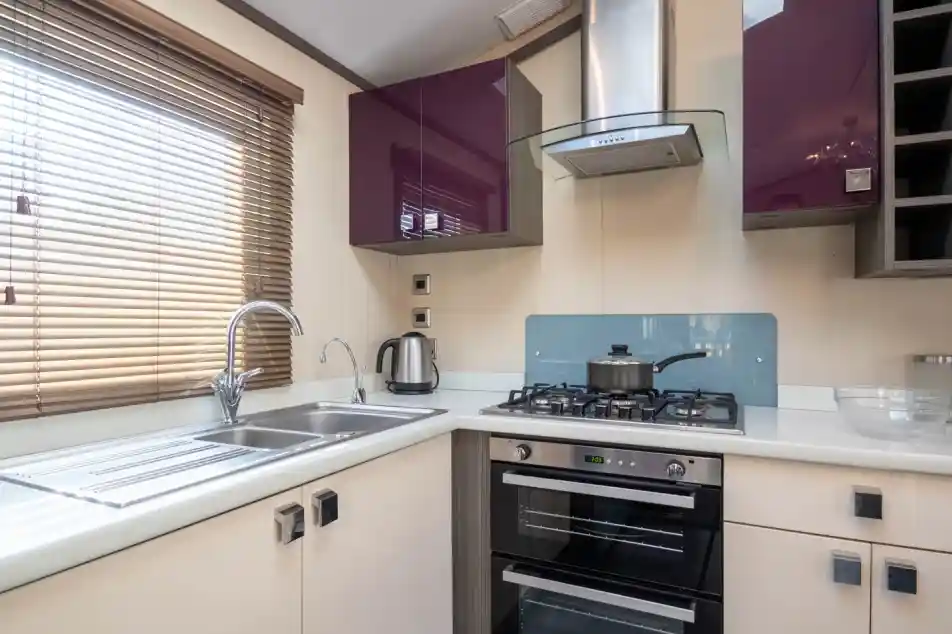 A bright small kitchen with purple and cream gloss cabinets, including an oven with gas hob, extractor hood and stainless steel one-and-a-half bowl sink unit, together with a window with blinds.