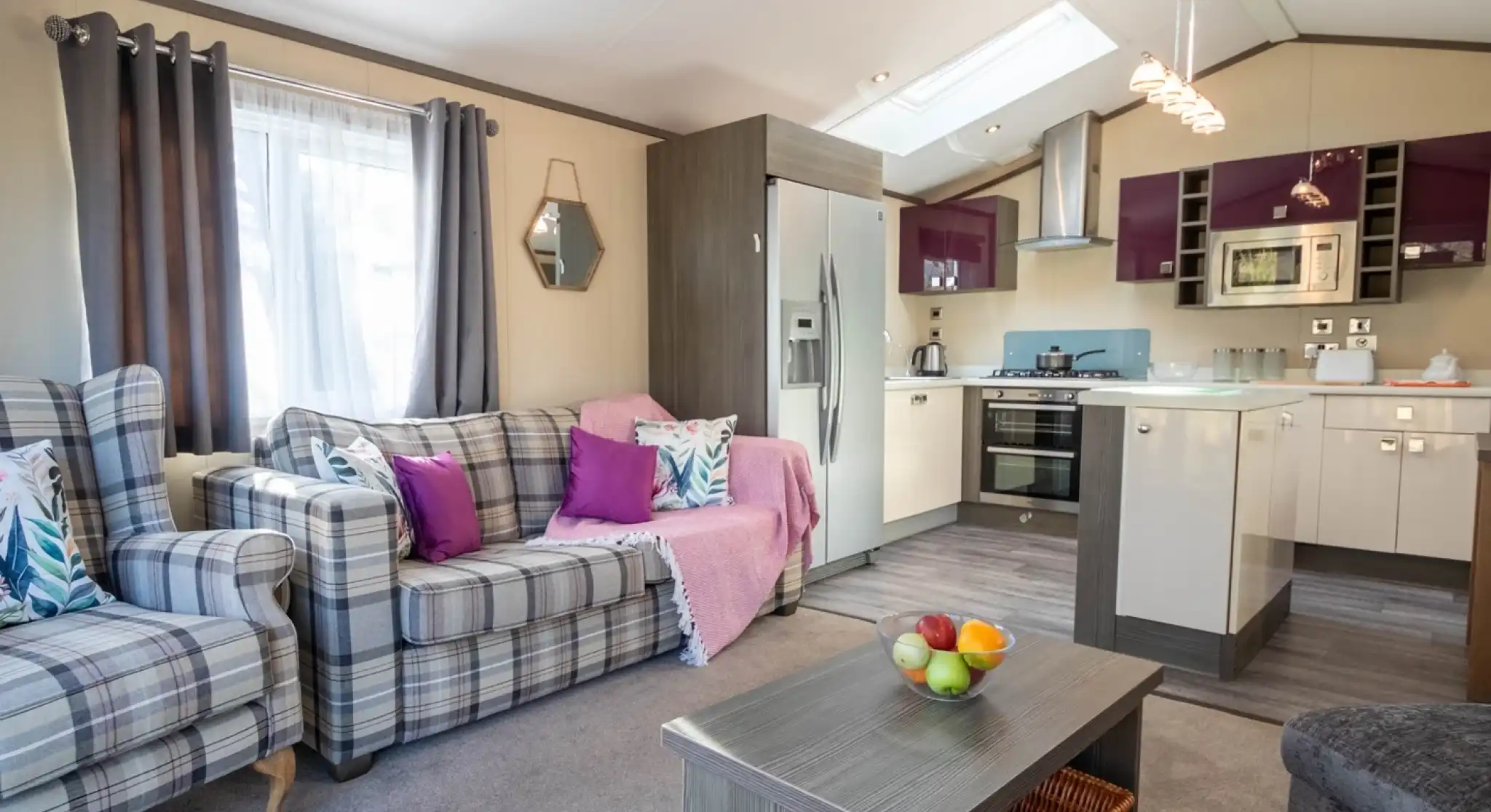 The image depicts a living room with free-standing patterned sofa and armchair with cushions in complementing bright colours, a coffee table with fruit bowl and large kitchen with gloss cabinetry in the background.