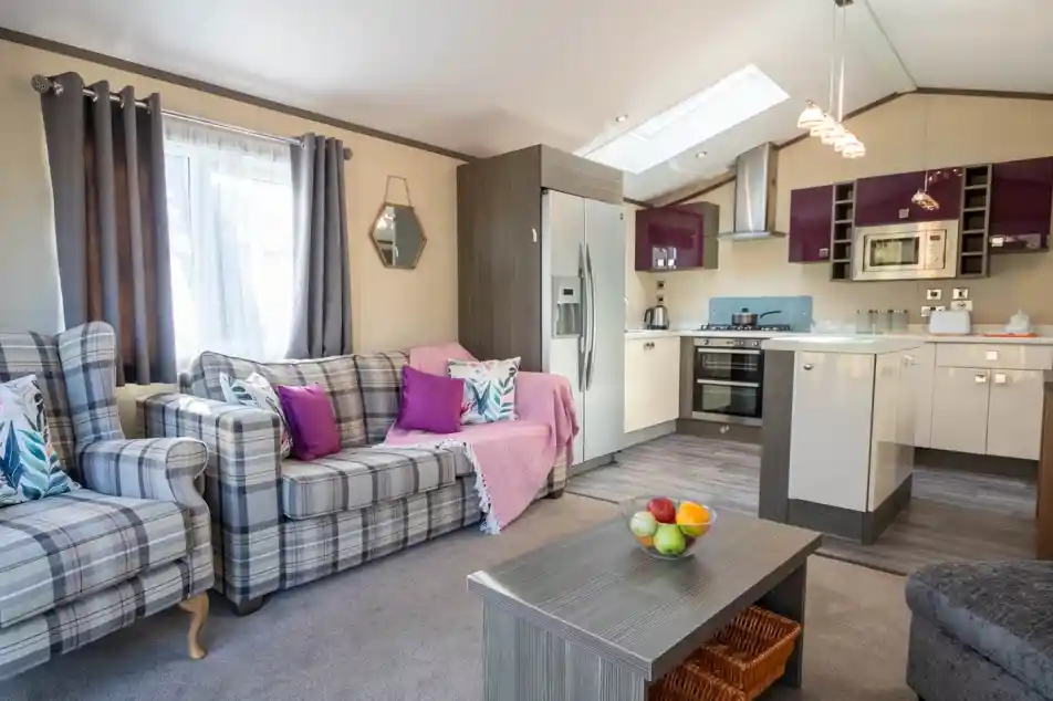 The image depicts a living room with free-standing patterned sofa and armchair with cushions in complementing bright colours, a coffee table with fruit bowl and large kitchen with gloss cabinetry in the background.