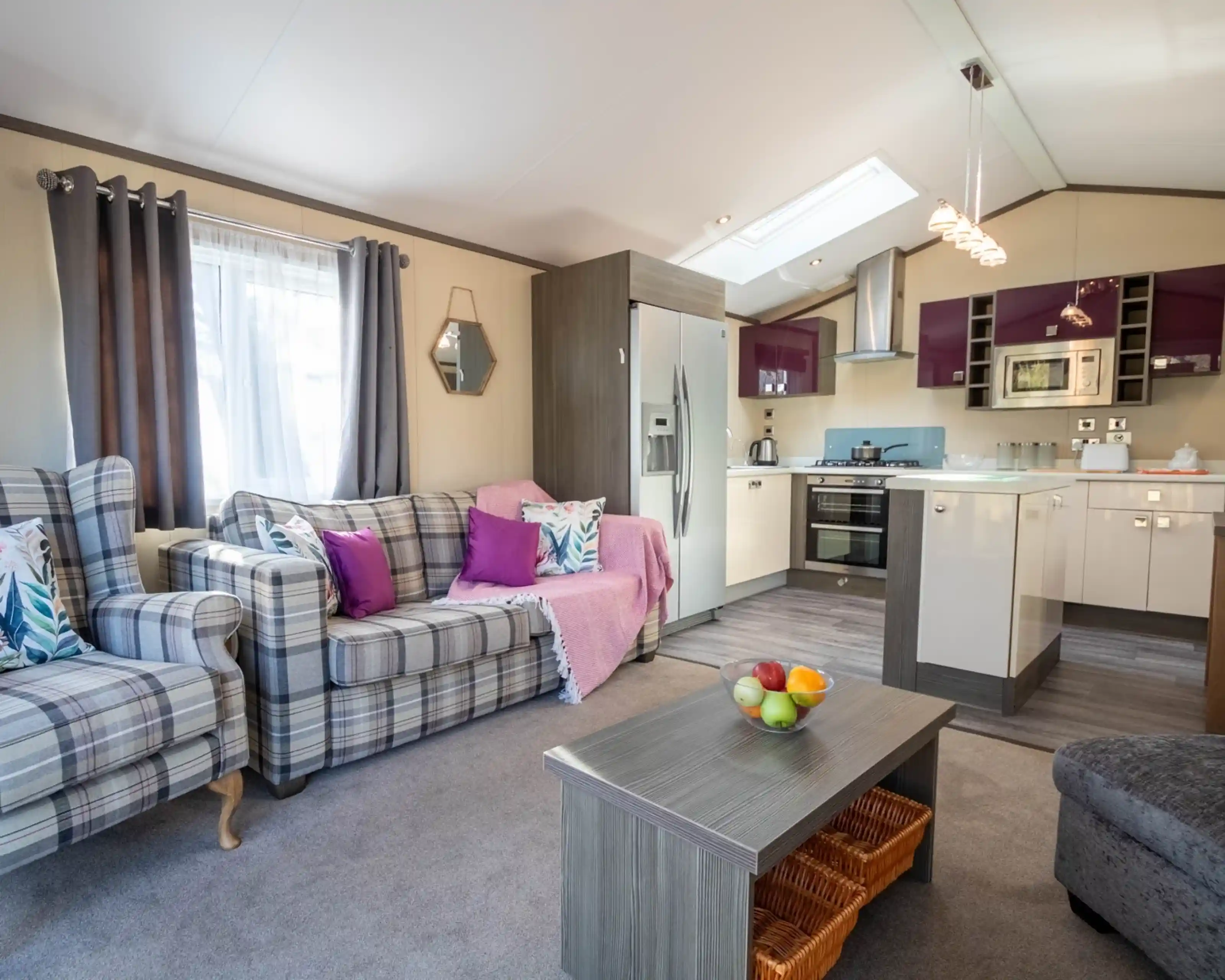 The image depicts a living room with free-standing patterned sofa and armchair with cushions in complementing bright colours, a coffee table with fruit bowl and large kitchen with gloss cabinetry in the background.