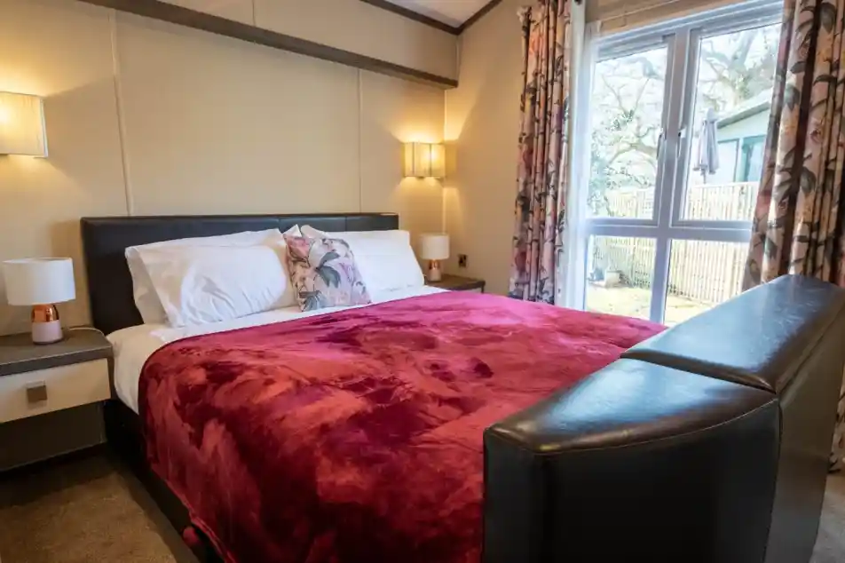 A cosy master bedroom with large double bed with bedside tables either side of the bed with lamps and wall lights. Floor length windows and curtains give an outlook outside.