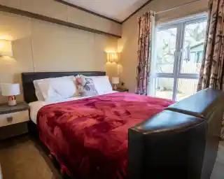 A cosy master bedroom with large double bed with bedside tables either side of the bed with lamps and wall lights. Floor length windows and curtains give an outlook outside.