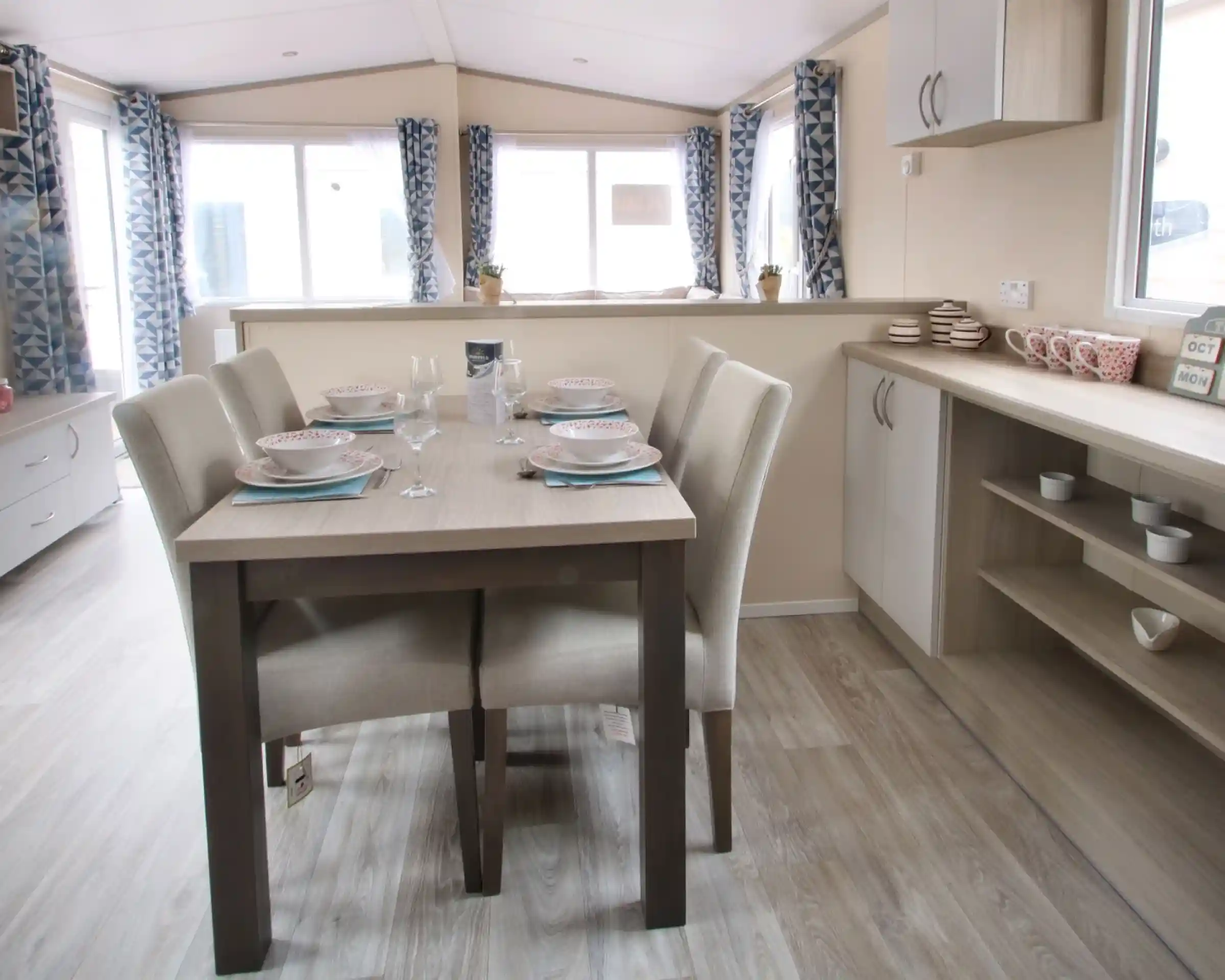 A dining table is positioned in a well-decorated room, surrounded by various pieces of furniture. The space features a window that allows natural light to enter, with cabinetry, countertops, and a sink visible nearby. The floor is adorned with suitable flooring for a kitchen or dining area, creating a cosy and inviting atmosphere.