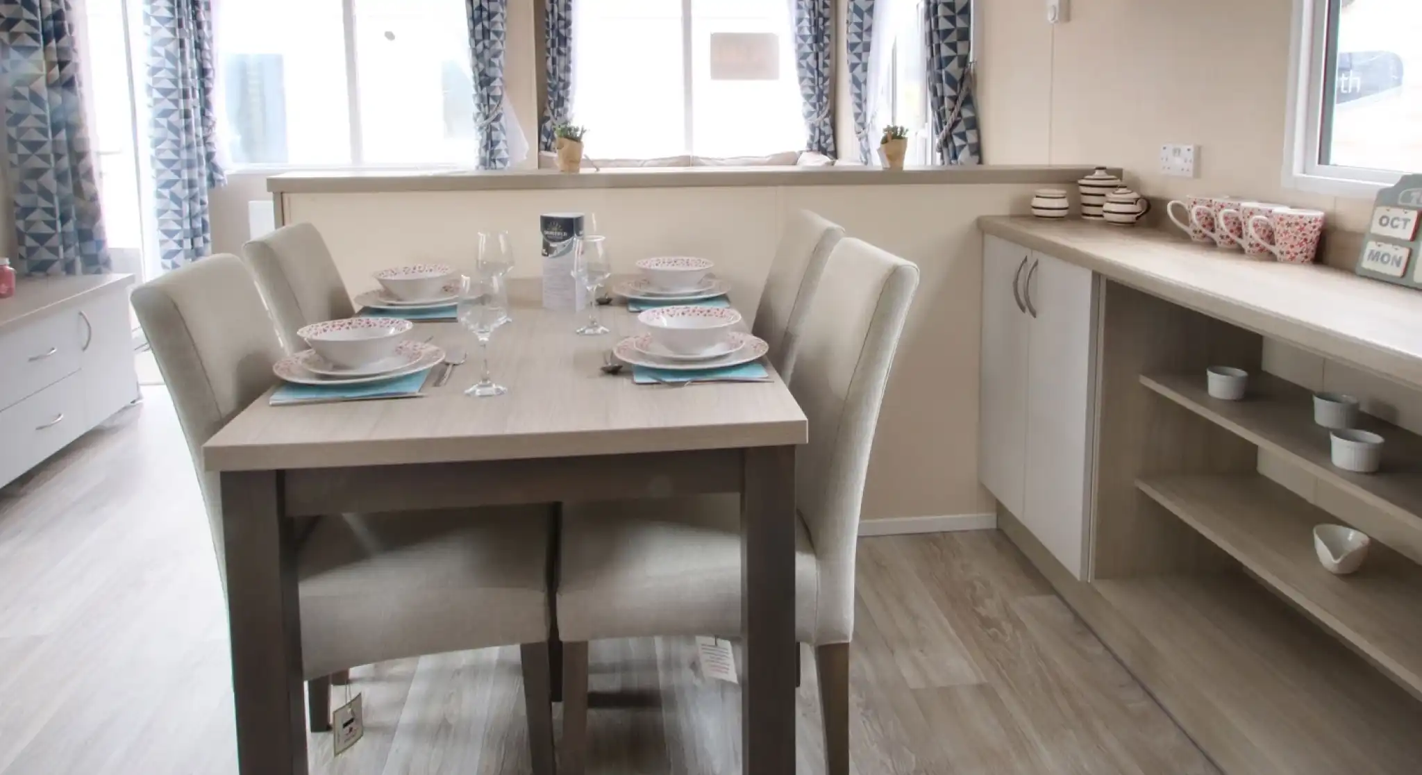 A dining table is positioned in a well-decorated room, surrounded by various pieces of furniture. The space features a window that allows natural light to enter, with cabinetry, countertops, and a sink visible nearby. The floor is adorned with suitable flooring for a kitchen or dining area, creating a cosy and inviting atmosphere.