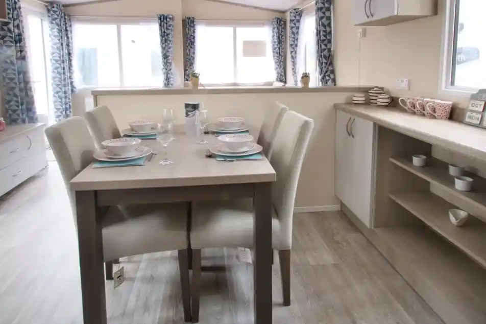 A dining table is positioned in a well-decorated room, surrounded by various pieces of furniture. The space features a window that allows natural light to enter, with cabinetry, countertops, and a sink visible nearby. The floor is adorned with suitable flooring for a kitchen or dining area, creating a cosy and inviting atmosphere.