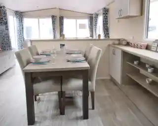 A dining table is positioned in a well-decorated room, surrounded by various pieces of furniture. The space features a window that allows natural light to enter, with cabinetry, countertops, and a sink visible nearby. The floor is adorned with suitable flooring for a kitchen or dining area, creating a cosy and inviting atmosphere.