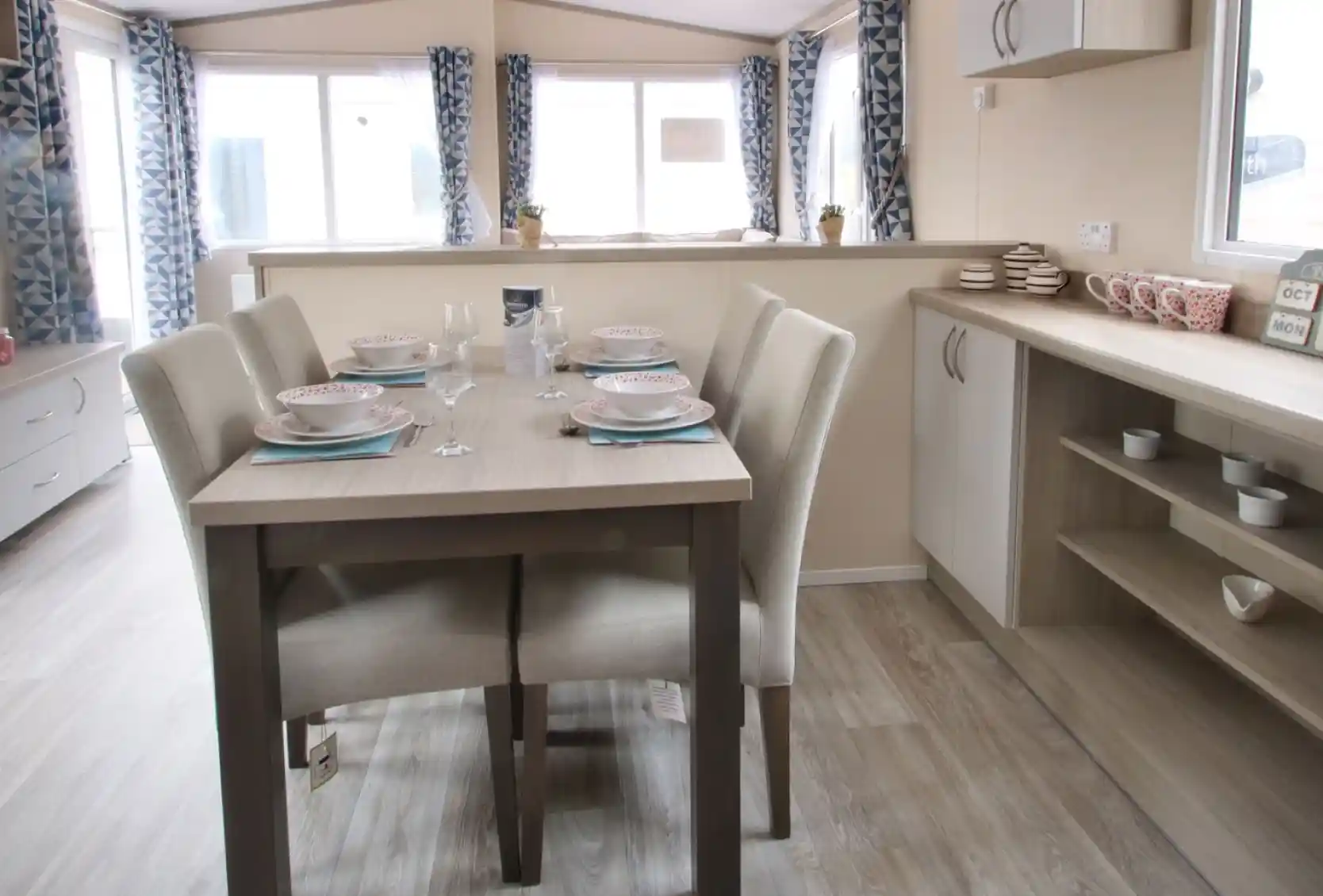 A dining table is positioned in a well-decorated room, surrounded by various pieces of furniture. The space features a window that allows natural light to enter, with cabinetry, countertops, and a sink visible nearby. The floor is adorned with suitable flooring for a kitchen or dining area, creating a cosy and inviting atmosphere.