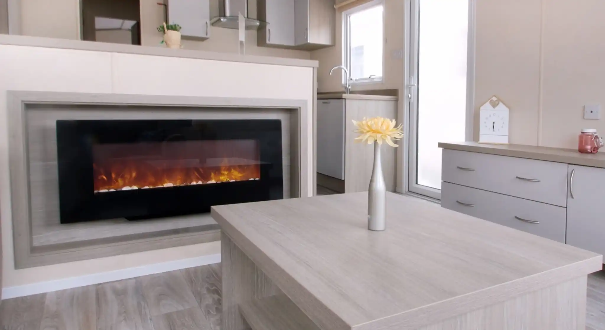 A modern living room with a wall-mounted electric fireplace, light wood furniture, a vase with yellow flowers on the table, and a small kitchen area in the background.