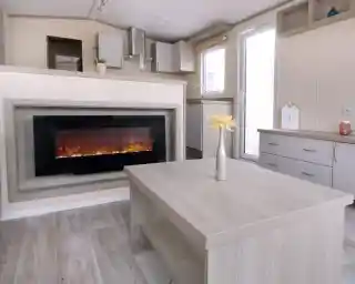 A modern living room with a wall-mounted electric fireplace, light wood furniture, a vase with yellow flowers on the table, and a small kitchen area in the background.