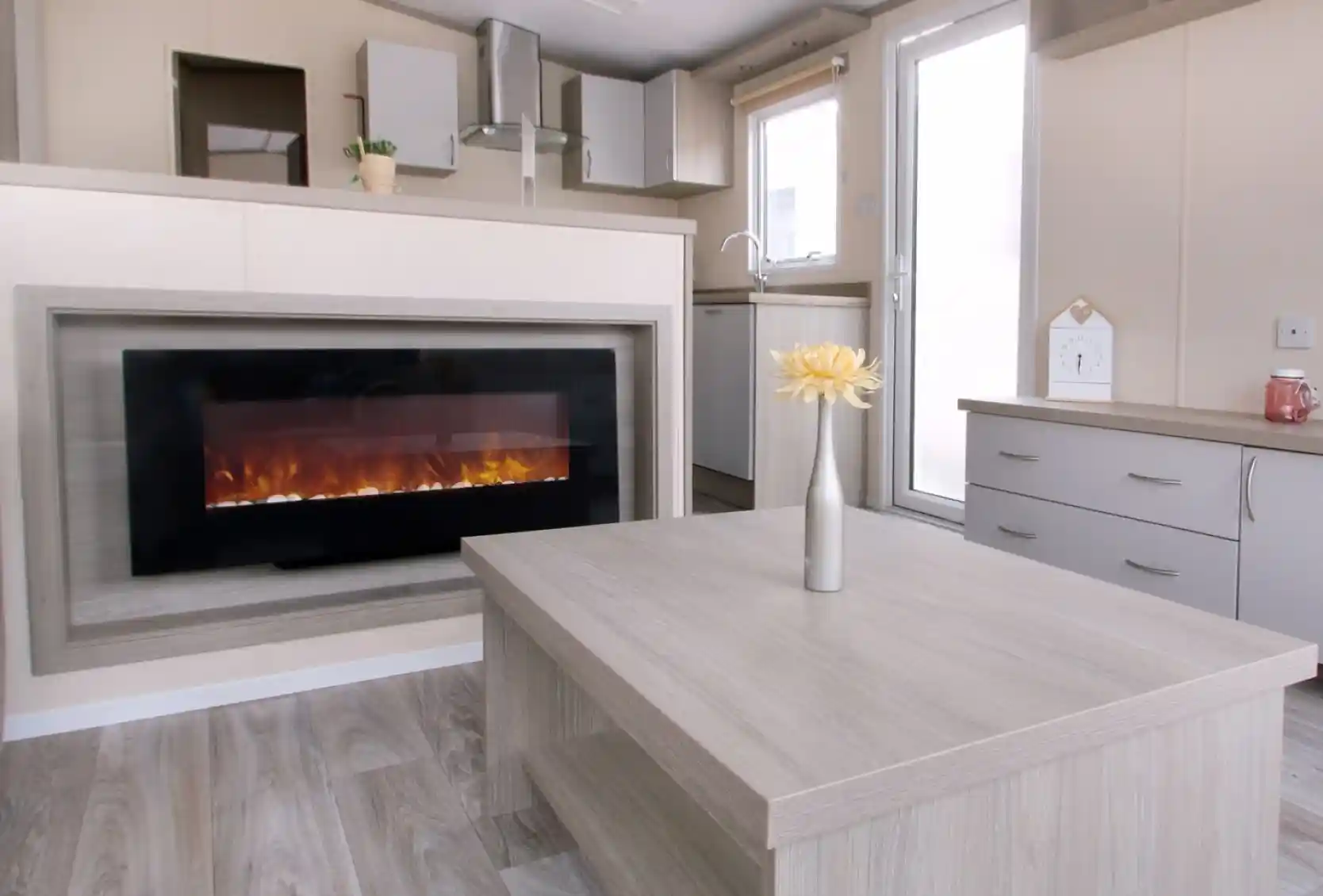 A modern living room with a wall-mounted electric fireplace, light wood furniture, a vase with yellow flowers on the table, and a small kitchen area in the background.
