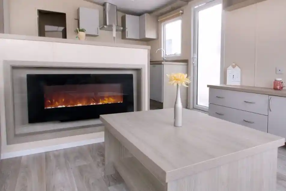 A modern living room with a wall-mounted electric fireplace, light wood furniture, a vase with yellow flowers on the table, and a small kitchen area in the background.