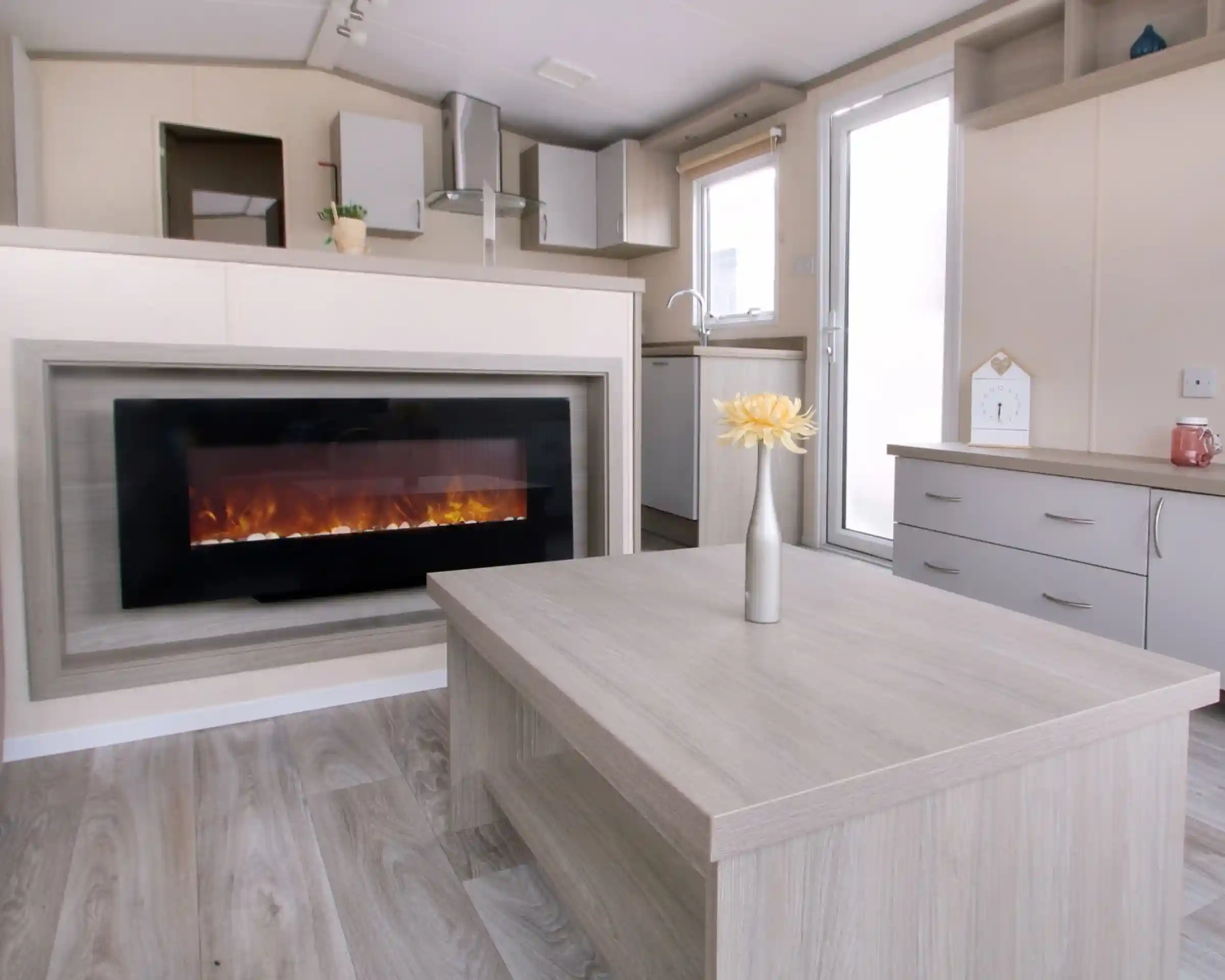 A modern living room with a wall-mounted electric fireplace, light wood furniture, a vase with yellow flowers on the table, and a small kitchen area in the background.