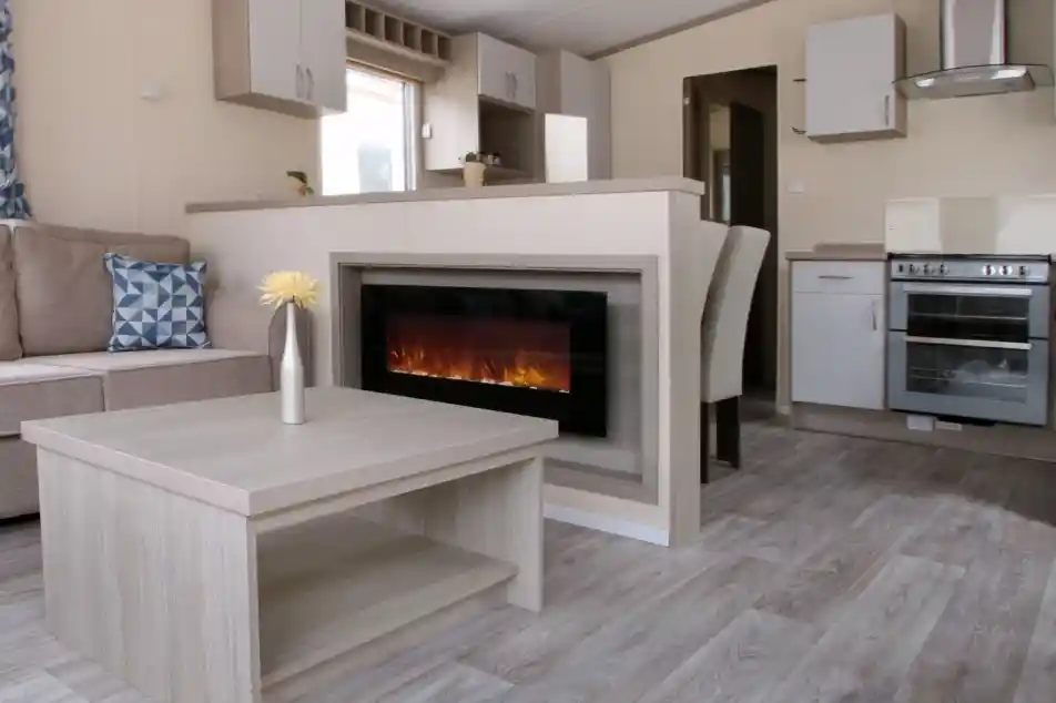 A cosy living room featuring a fireplace, with an adjoining kitchen area. The space is designed with wood and laminate flooring, showcasing a comfortable couch and cabinetry. The decor reflects a warm and inviting home atmosphere.