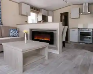 A cosy living room featuring a fireplace, with an adjoining kitchen area. The space is designed with wood and laminate flooring, showcasing a comfortable couch and cabinetry. The decor reflects a warm and inviting home atmosphere.