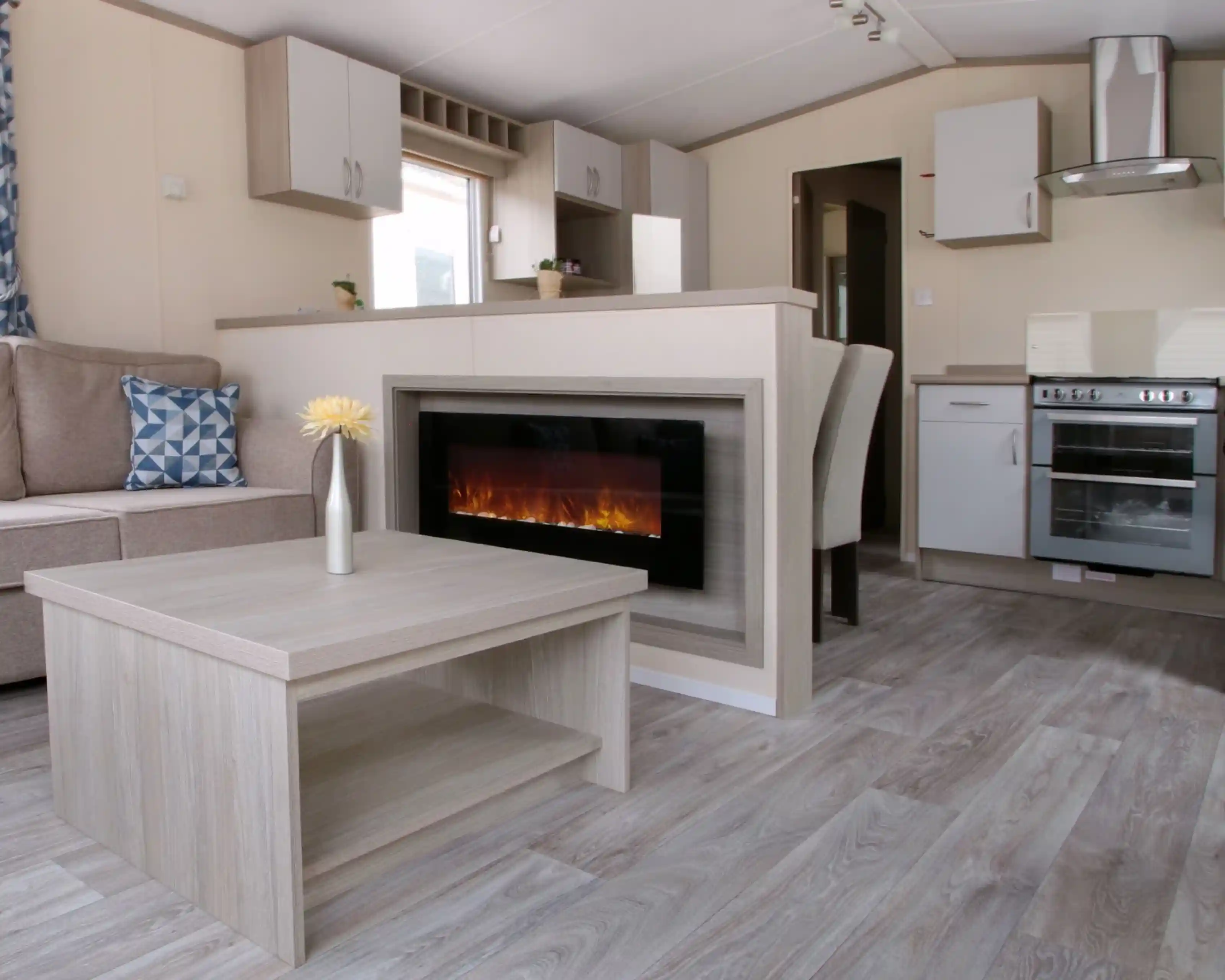 A cosy living room featuring a fireplace, with an adjoining kitchen area. The space is designed with wood and laminate flooring, showcasing a comfortable couch and cabinetry. The decor reflects a warm and inviting home atmosphere.