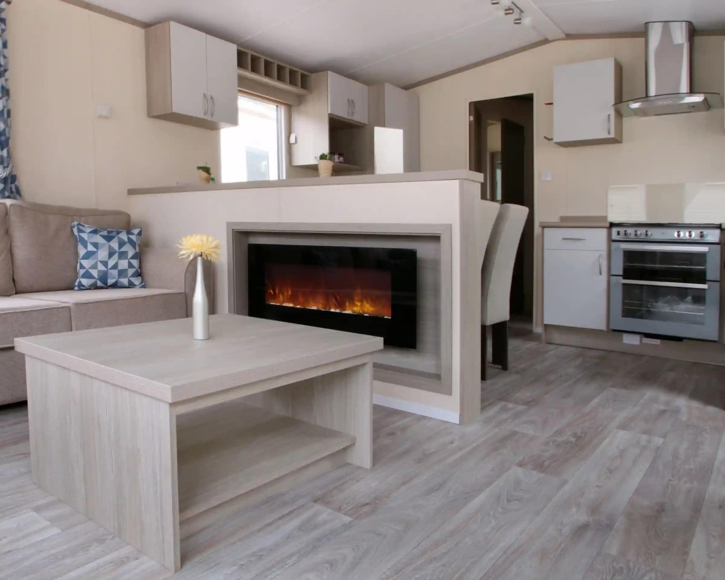 A cosy living room featuring a fireplace, with an adjoining kitchen area. The space is designed with wood and laminate flooring, showcasing a comfortable couch and cabinetry. The decor reflects a warm and inviting home atmosphere.