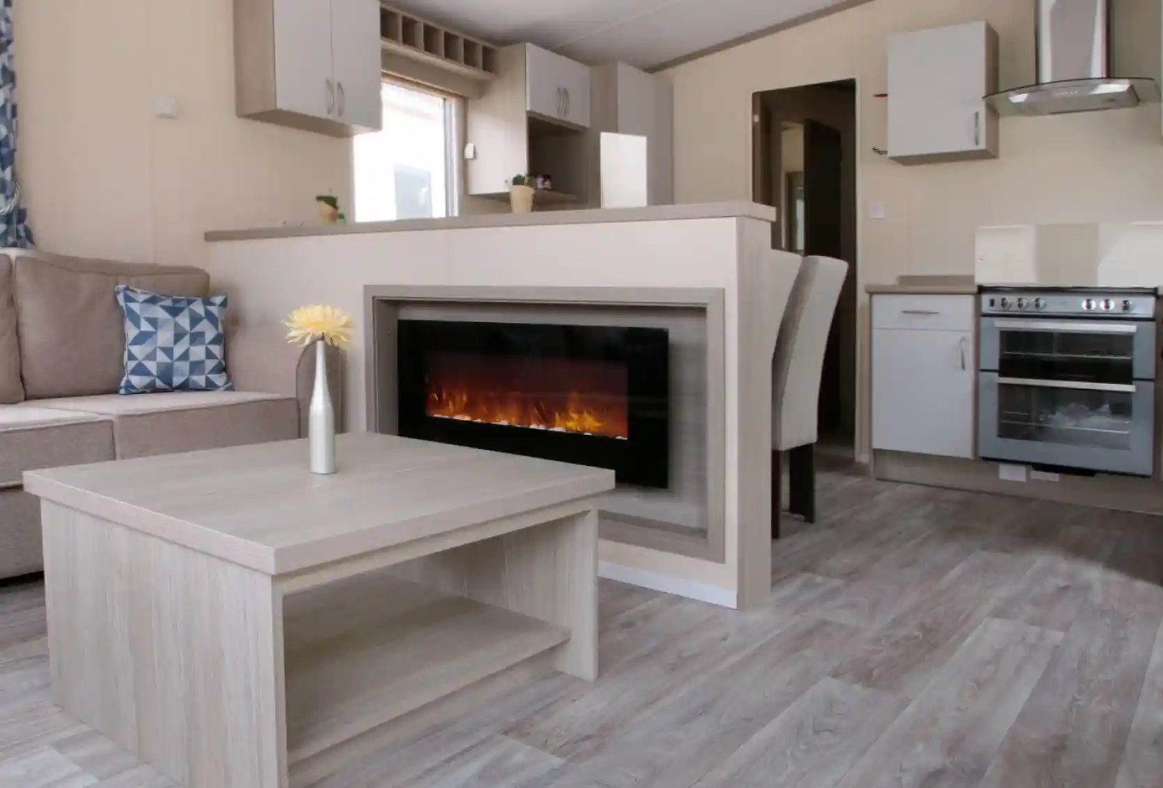 A cosy living room featuring a fireplace, with an adjoining kitchen area. The space is designed with wood and laminate flooring, showcasing a comfortable couch and cabinetry. The decor reflects a warm and inviting home atmosphere.