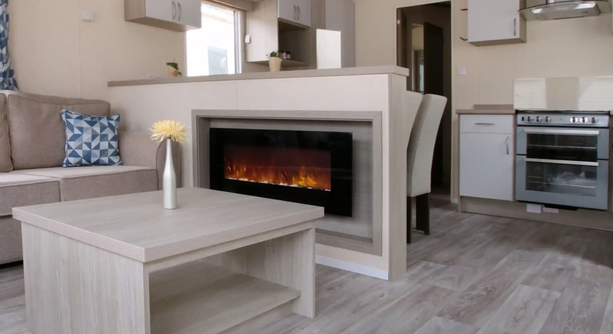 A cosy living room featuring a fireplace, with an adjoining kitchen area. The space is designed with wood and laminate flooring, showcasing a comfortable couch and cabinetry. The decor reflects a warm and inviting home atmosphere.