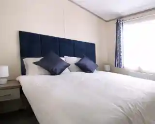 A neatly made bed with multiple pillows and a blue headboard, set in a stylish bedroom featuring soft linens and decorative cushions over a mattress. The bed is complemented by a nightstand and various interior design elements, including window treatments and lampshades.