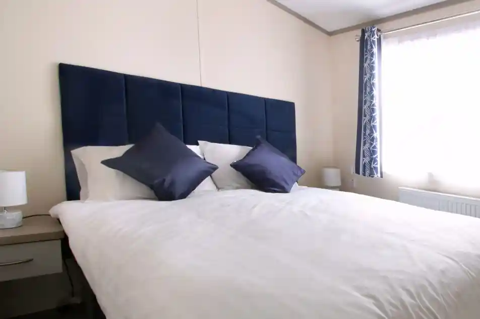 A neatly made bed with multiple pillows and a blue headboard, set in a stylish bedroom featuring soft linens and decorative cushions over a mattress. The bed is complemented by a nightstand and various interior design elements, including window treatments and lampshades.