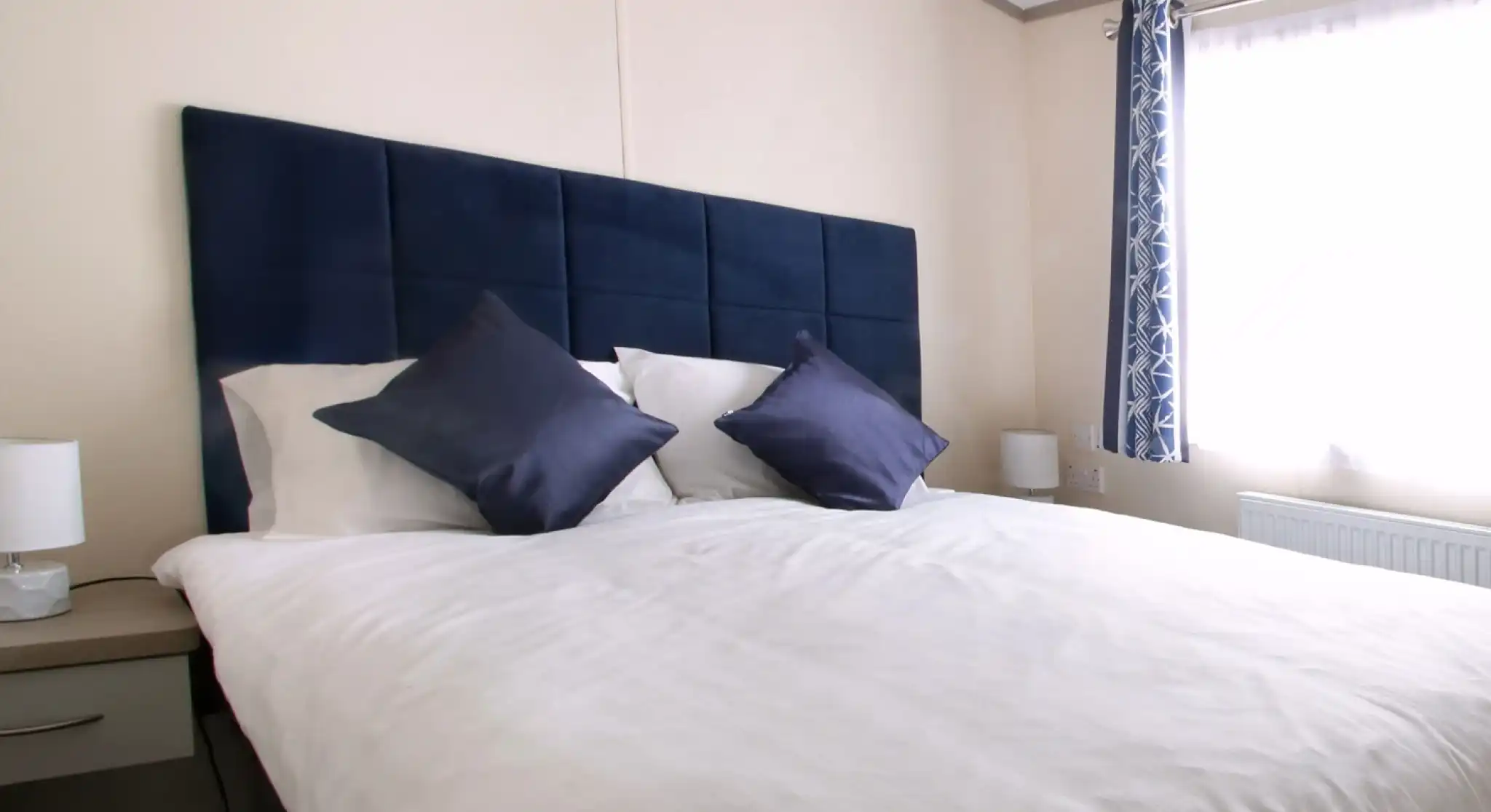 A neatly made bed with multiple pillows and a blue headboard, set in a stylish bedroom featuring soft linens and decorative cushions over a mattress. The bed is complemented by a nightstand and various interior design elements, including window treatments and lampshades.