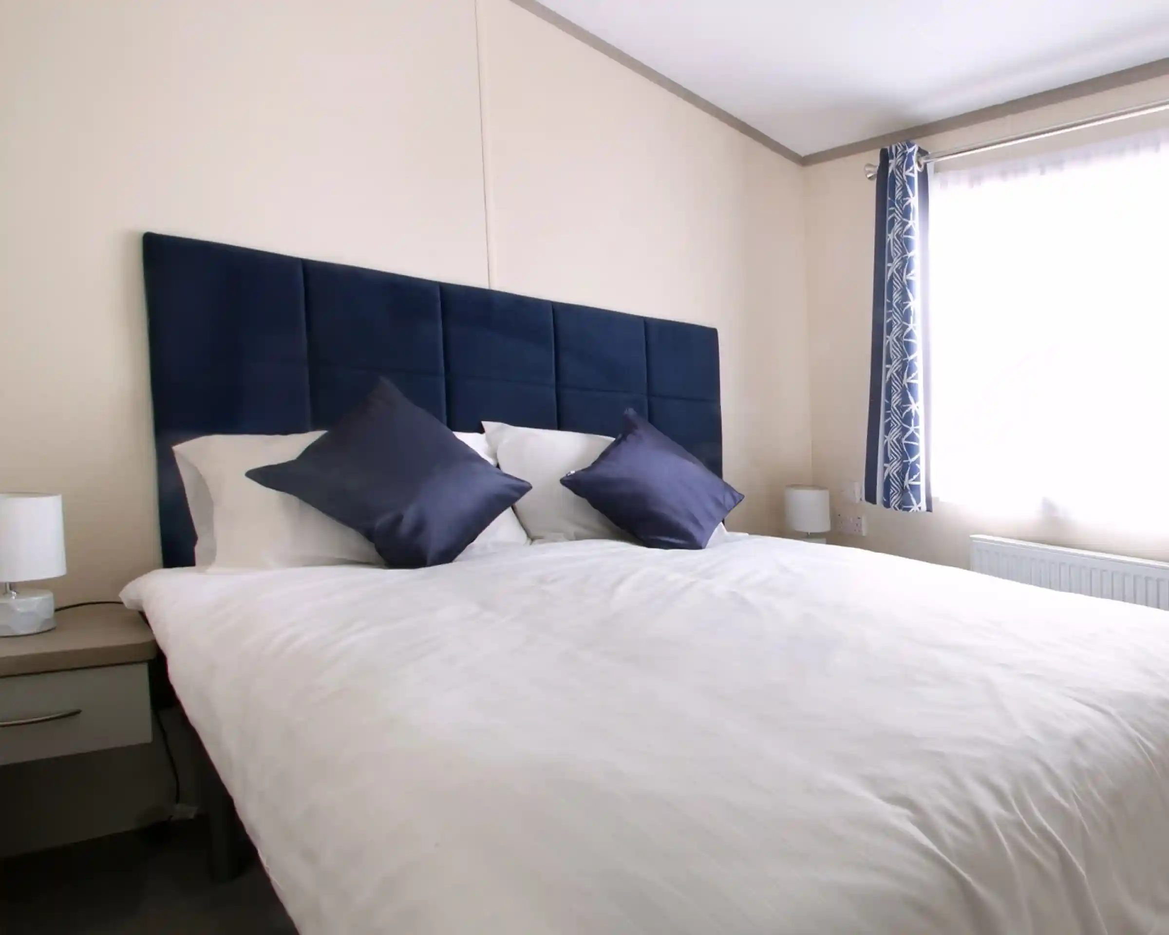 A neatly made bed with multiple pillows and a blue headboard, set in a stylish bedroom featuring soft linens and decorative cushions over a mattress. The bed is complemented by a nightstand and various interior design elements, including window treatments and lampshades.