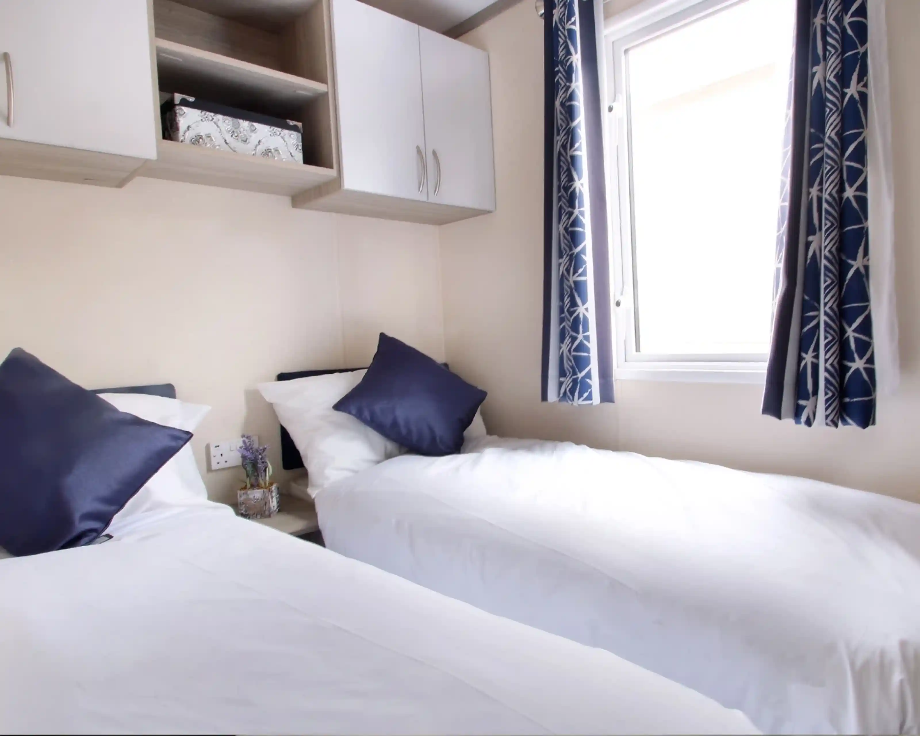 Two beds with beige bedding in a well-decorated room. Each bed features a headboard and decorative throw pillows, alongside nightstands. Natural light filters through sheer curtains, highlighting the stylish interior design elements like cushions.