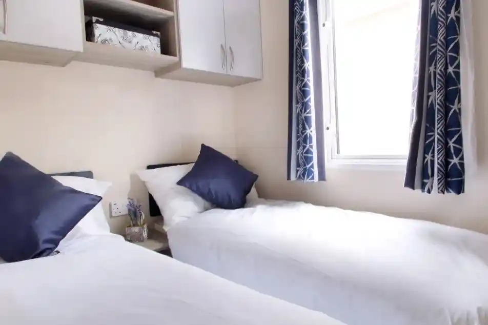 Two beds with beige bedding in a well-decorated room. Each bed features a headboard and decorative throw pillows, alongside nightstands. Natural light filters through sheer curtains, highlighting the stylish interior design elements like cushions.