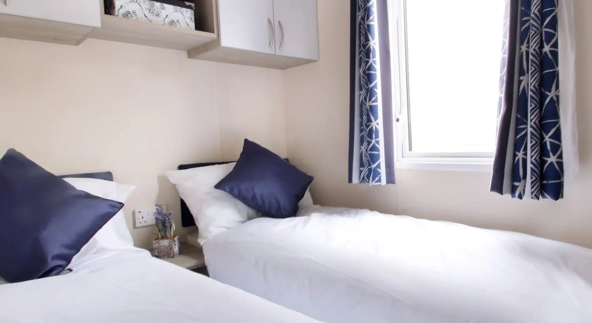 Two beds with beige bedding in a well-decorated room. Each bed features a headboard and decorative throw pillows, alongside nightstands. Natural light filters through sheer curtains, highlighting the stylish interior design elements like cushions.