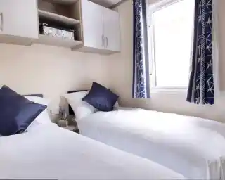 Two beds with beige bedding in a well-decorated room. Each bed features a headboard and decorative throw pillows, alongside nightstands. Natural light filters through sheer curtains, highlighting the stylish interior design elements like cushions.