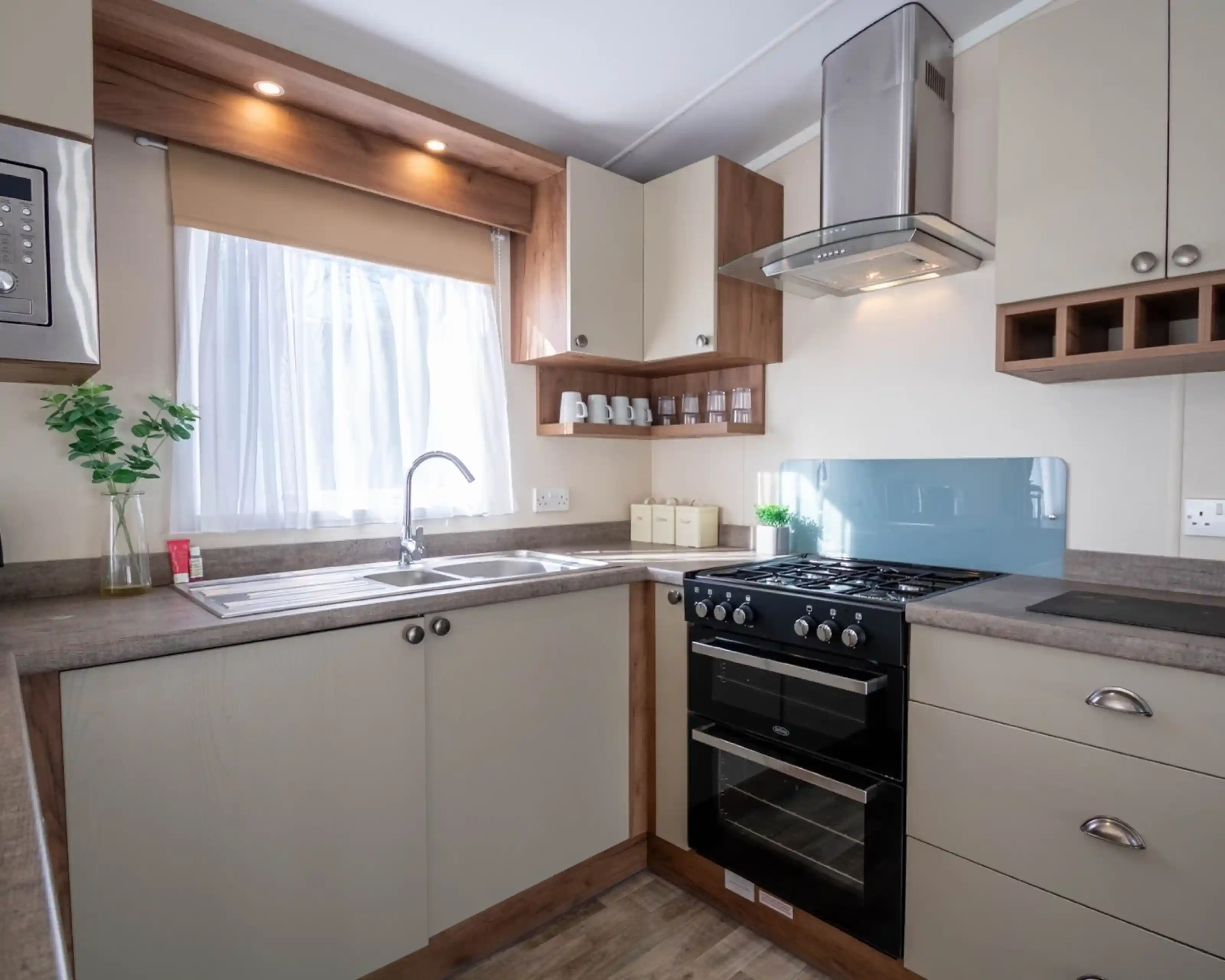 A kitchen with cream cabinets, an oven with gab hob and extractor over, and stainless steel sink unit with window above.
