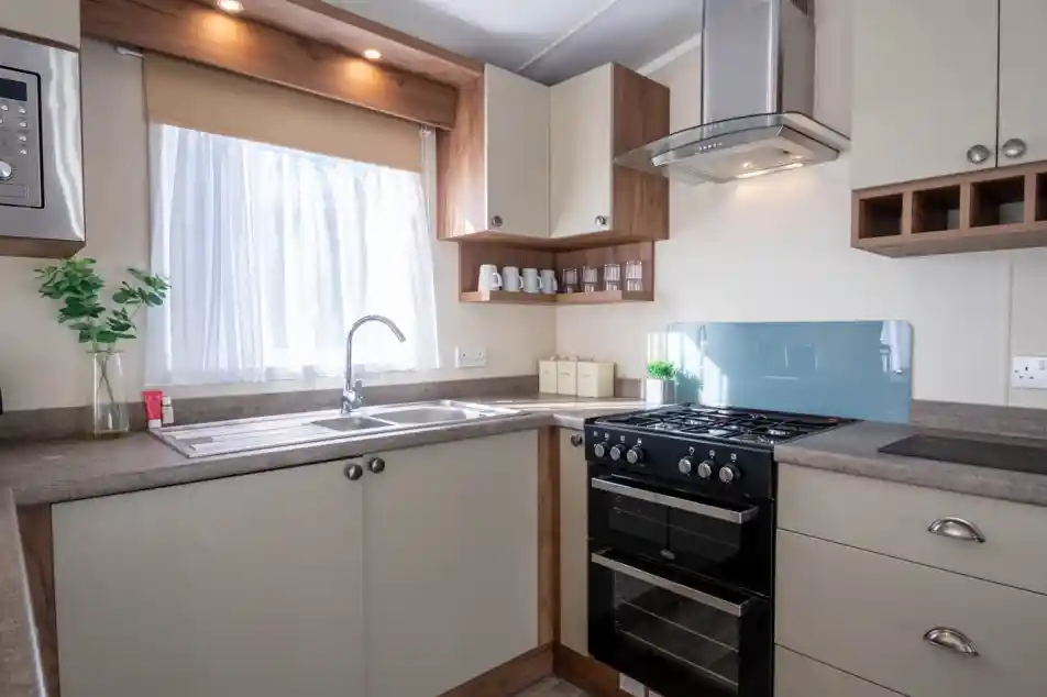 A kitchen with cream cabinets, an oven with gab hob and extractor over, and stainless steel sink unit with window above.