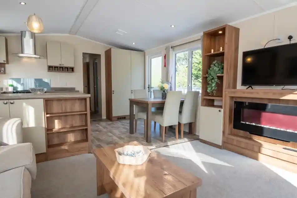 A lounge with coffee table, electric fireplace and TV, with kitchen and dining room in the background.