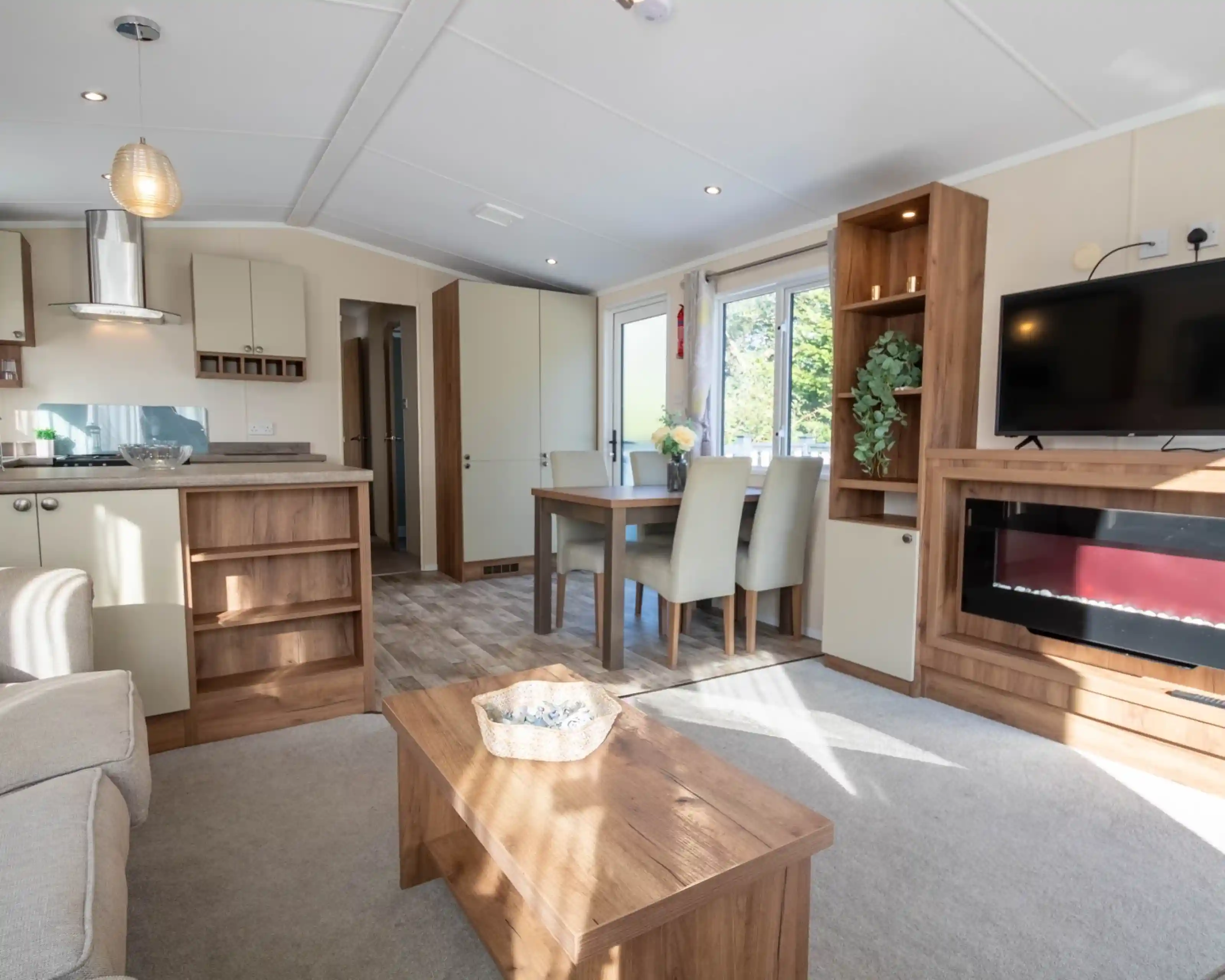 A lounge with coffee table, electric fireplace and TV, with kitchen and dining room in the background.