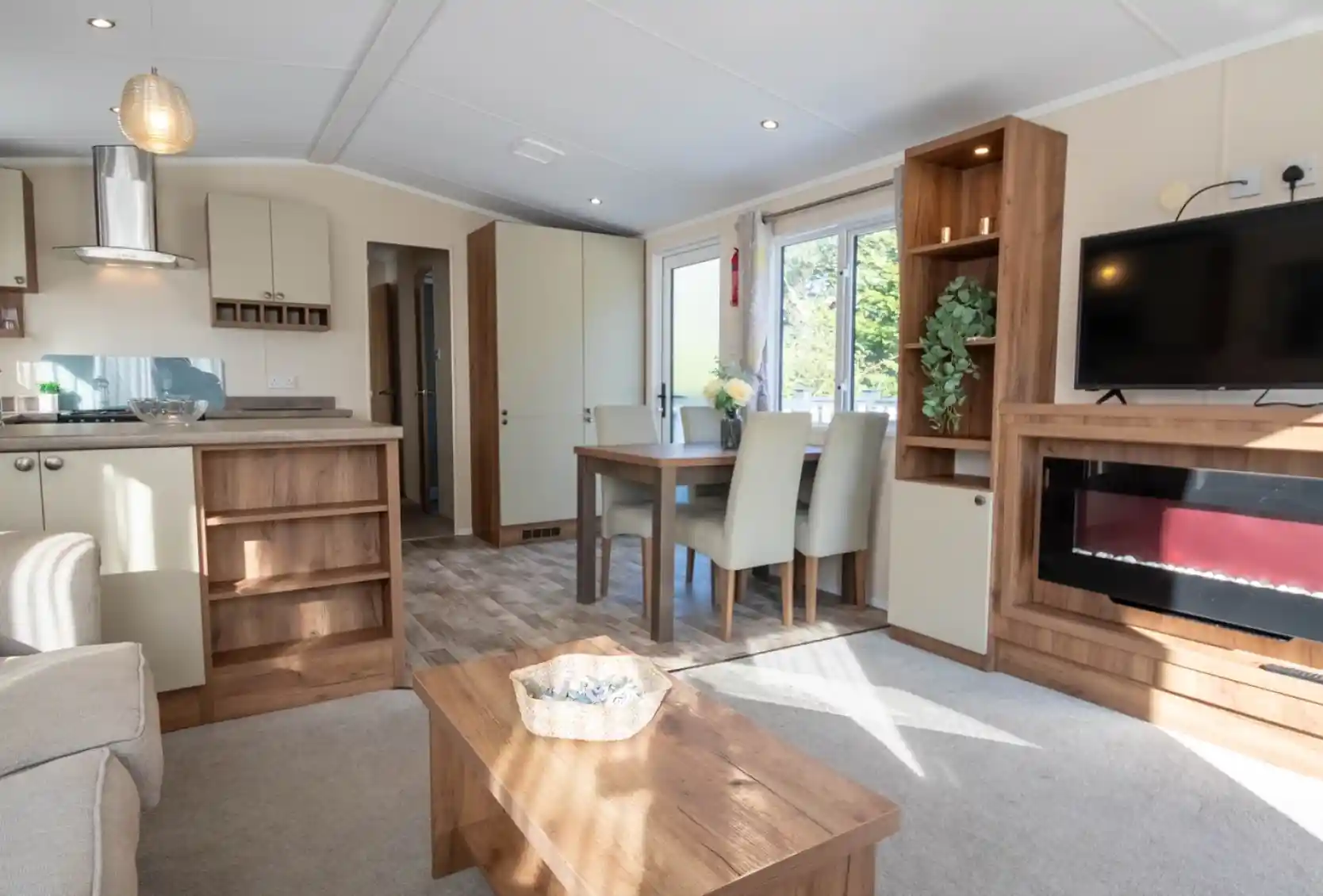 A lounge with coffee table, electric fireplace and TV, with kitchen and dining room in the background.