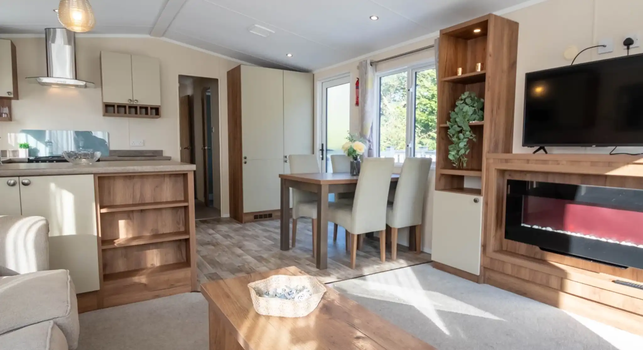 A lounge with coffee table, electric fireplace and TV, with kitchen and dining room in the background.