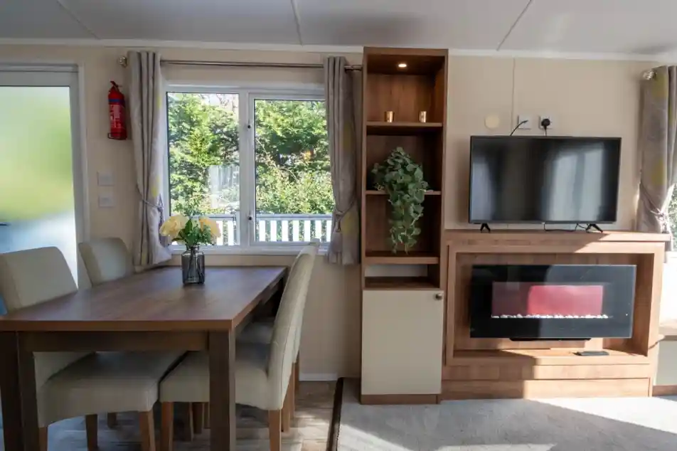 A dining area with dining table and chairs leading into a lounge with feature electric fireplace set into cabinets with a TV above.