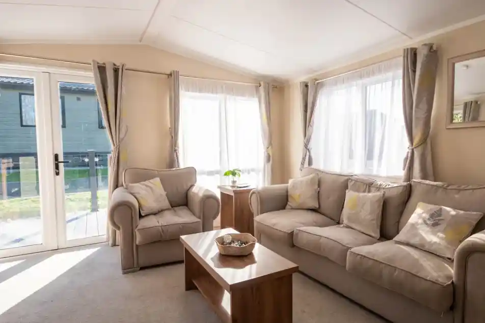 A lounge with three seater comfortable sofa and armchair with coffee table and patio doors and windows giving an outlook onto the decking.