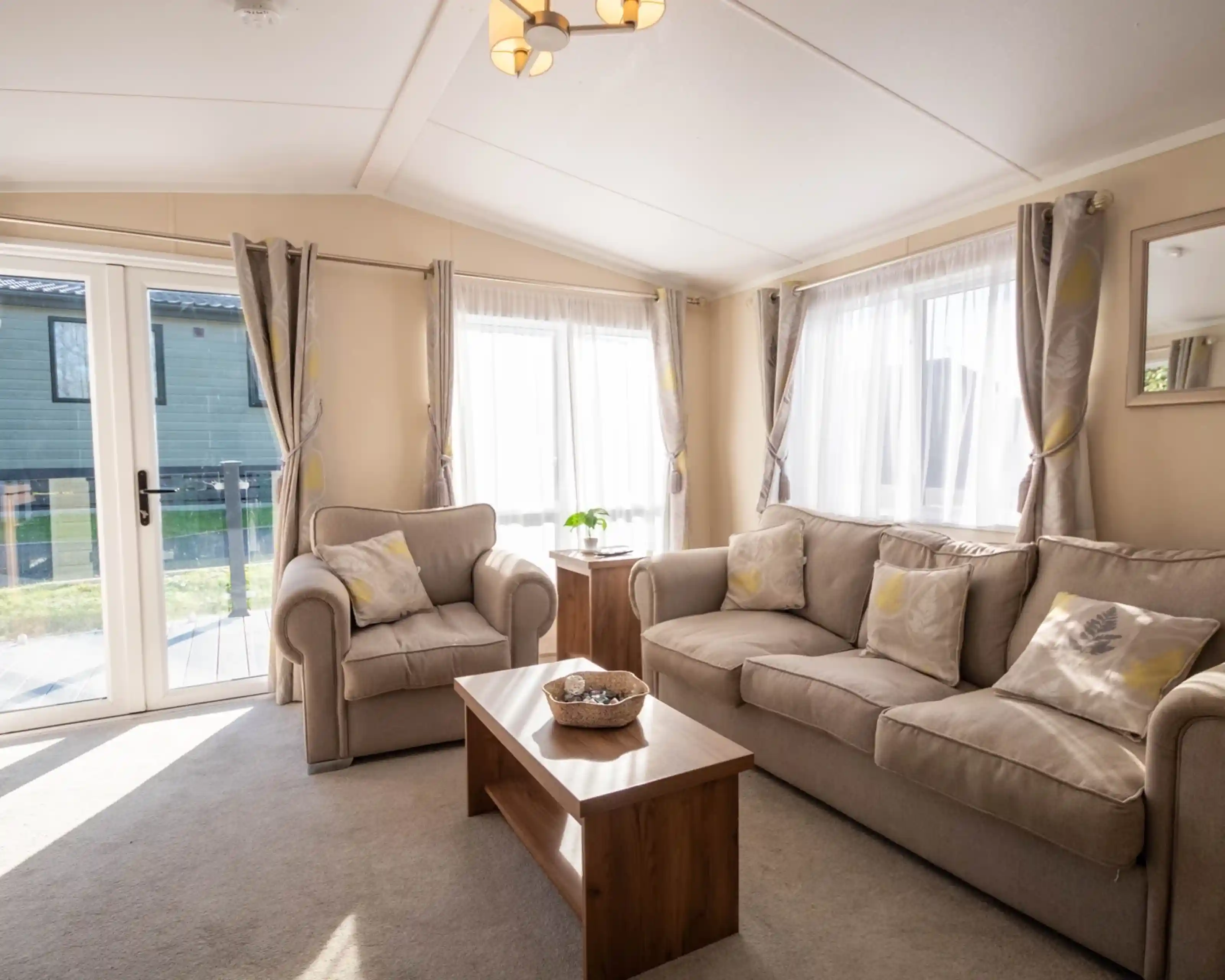 A lounge with three seater comfortable sofa and armchair with coffee table and patio doors and windows giving an outlook onto the decking.