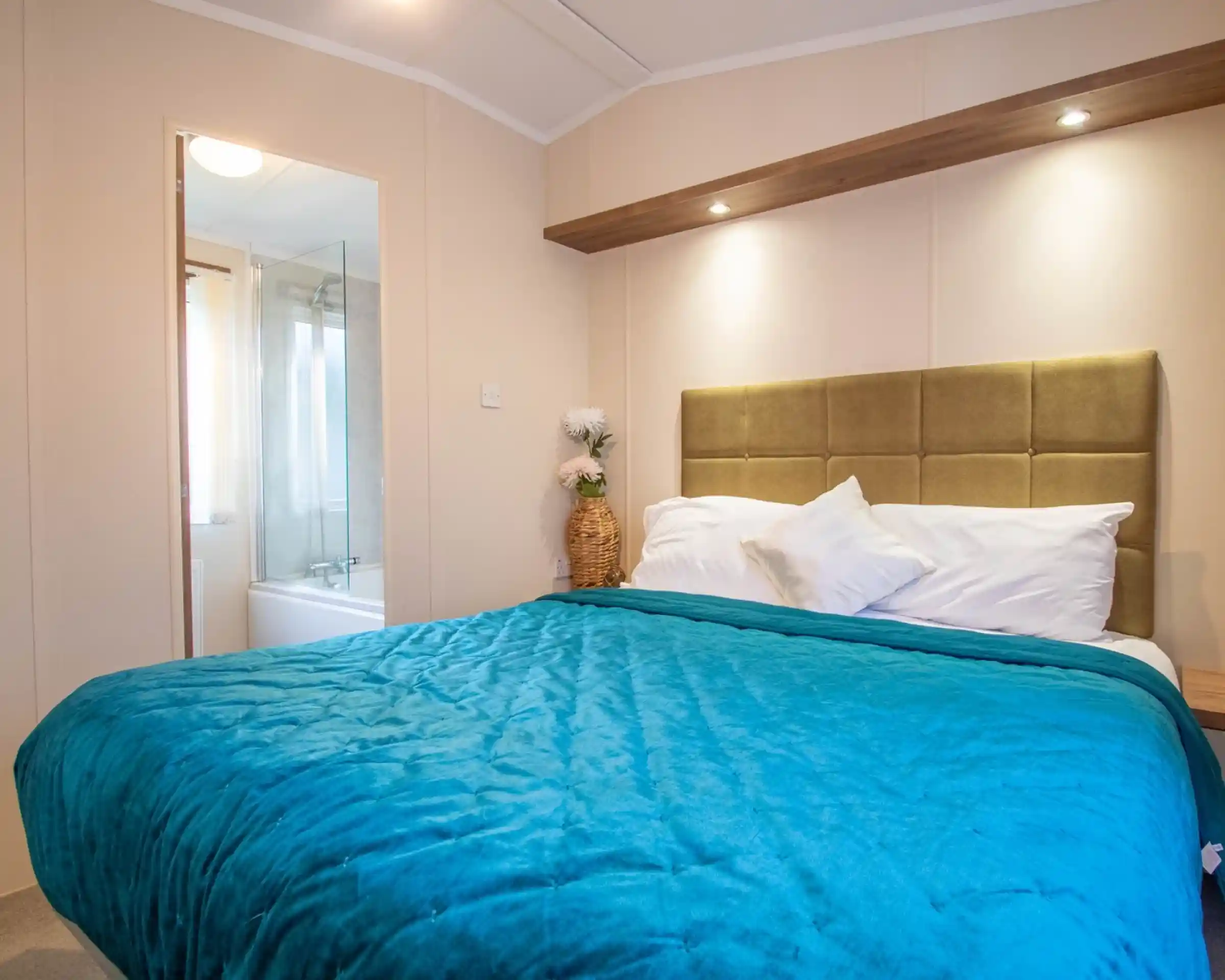 A master bedroom with a double bed with blue throw and gold plush headboard. There is an en suite bathroom with bath with shower over in the background.