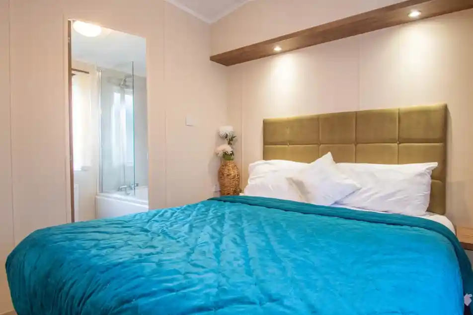 A master bedroom with a double bed with blue throw and gold plush headboard. There is an en suite bathroom with bath with shower over in the background.