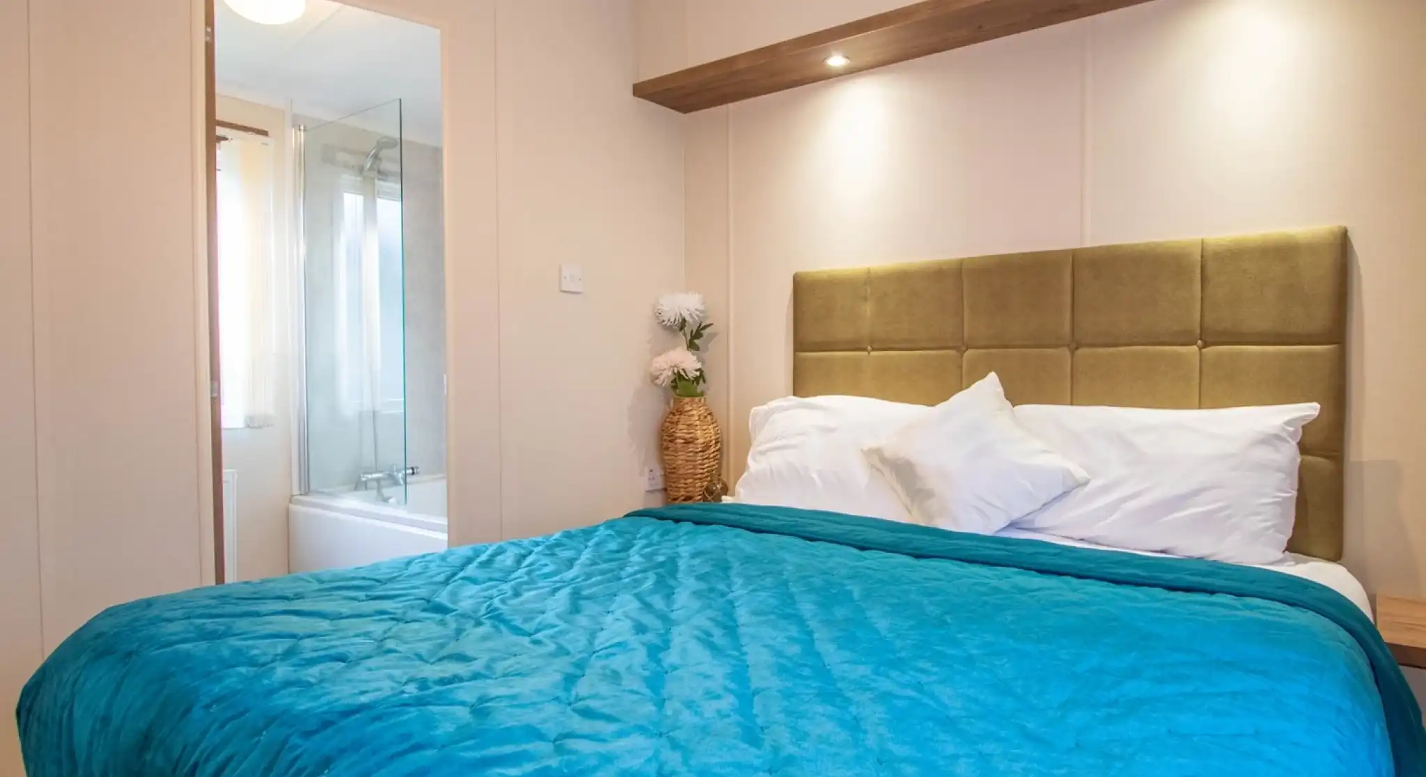 A master bedroom with a double bed with blue throw and gold plush headboard. There is an en suite bathroom with bath with shower over in the background.