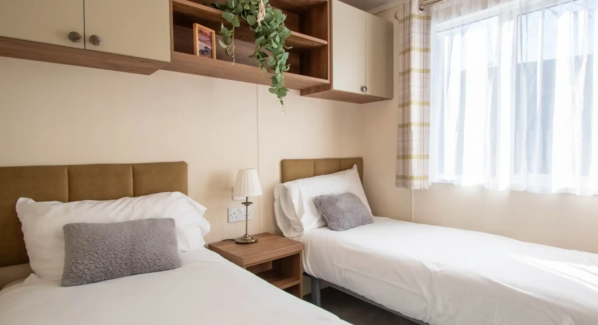 A bedroom featuring two beds with fold headboards and crisp white linen, overhead storage and a bedside table. There is a window providing natural light.