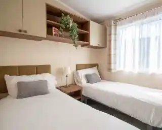 A bedroom featuring two beds with fold headboards and crisp white linen, overhead storage and a bedside table. There is a window providing natural light.
