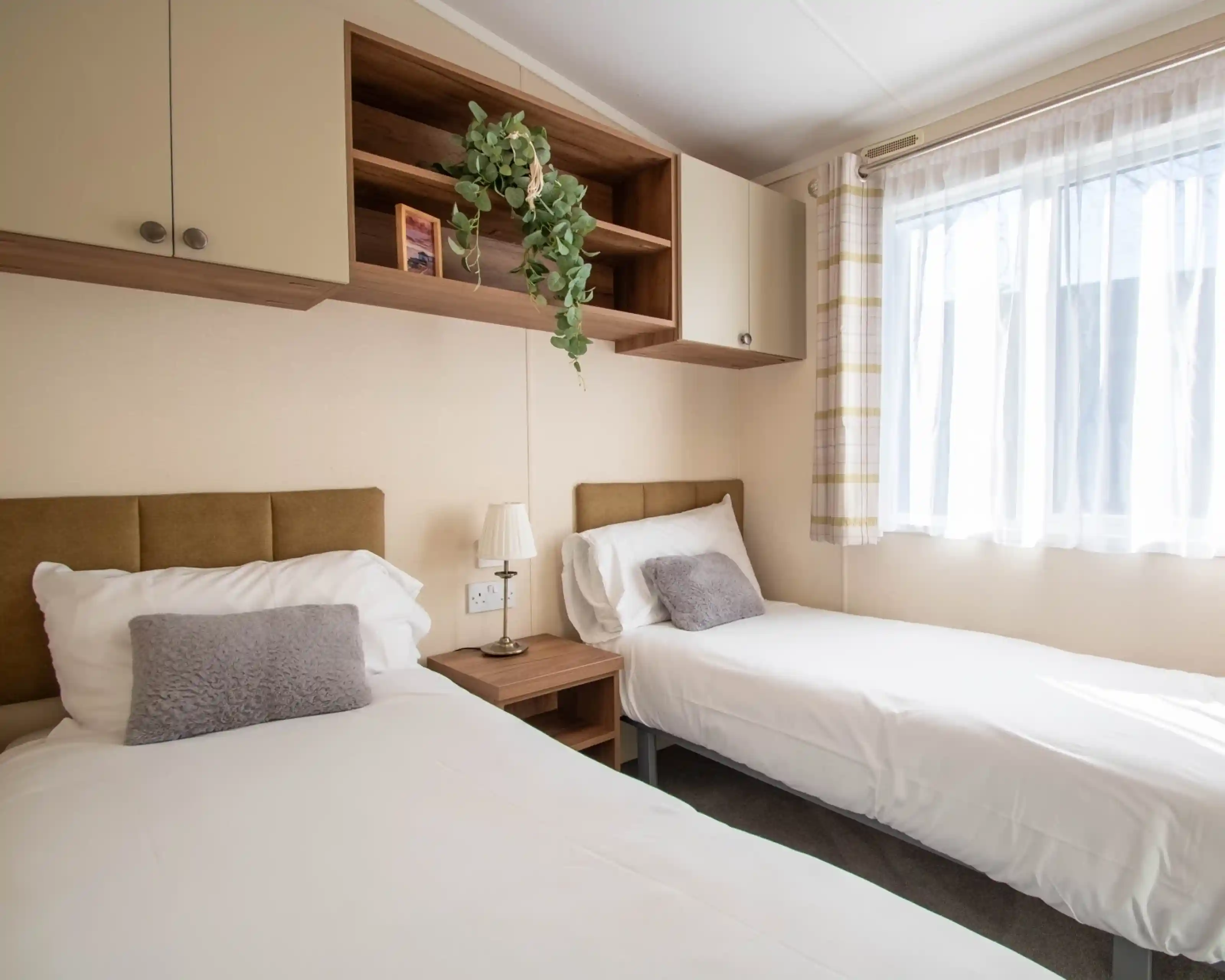 A bedroom featuring two beds with fold headboards and crisp white linen, overhead storage and a bedside table. There is a window providing natural light.